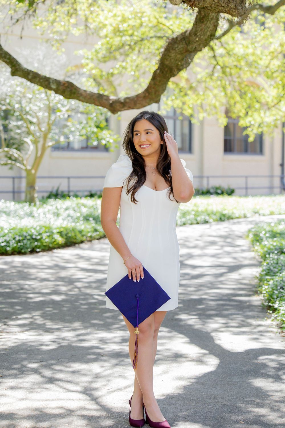 A graduate in a white dress stands on a tree-lined path holding a blue diploma folder while smiling outdoors.