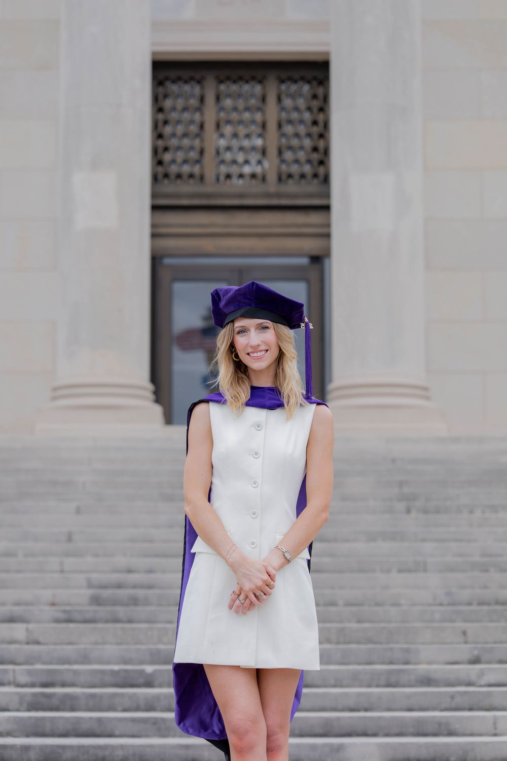 Graduate poses on stone steps wearing purple and white commencement regalia and graduation cap.