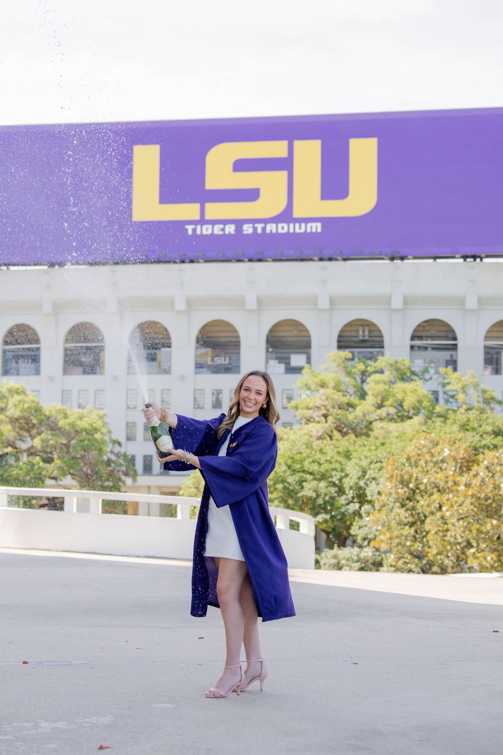 A graduate in a purple gown stands posing outside LSU Tiger Stadium with athletic signage visible above.