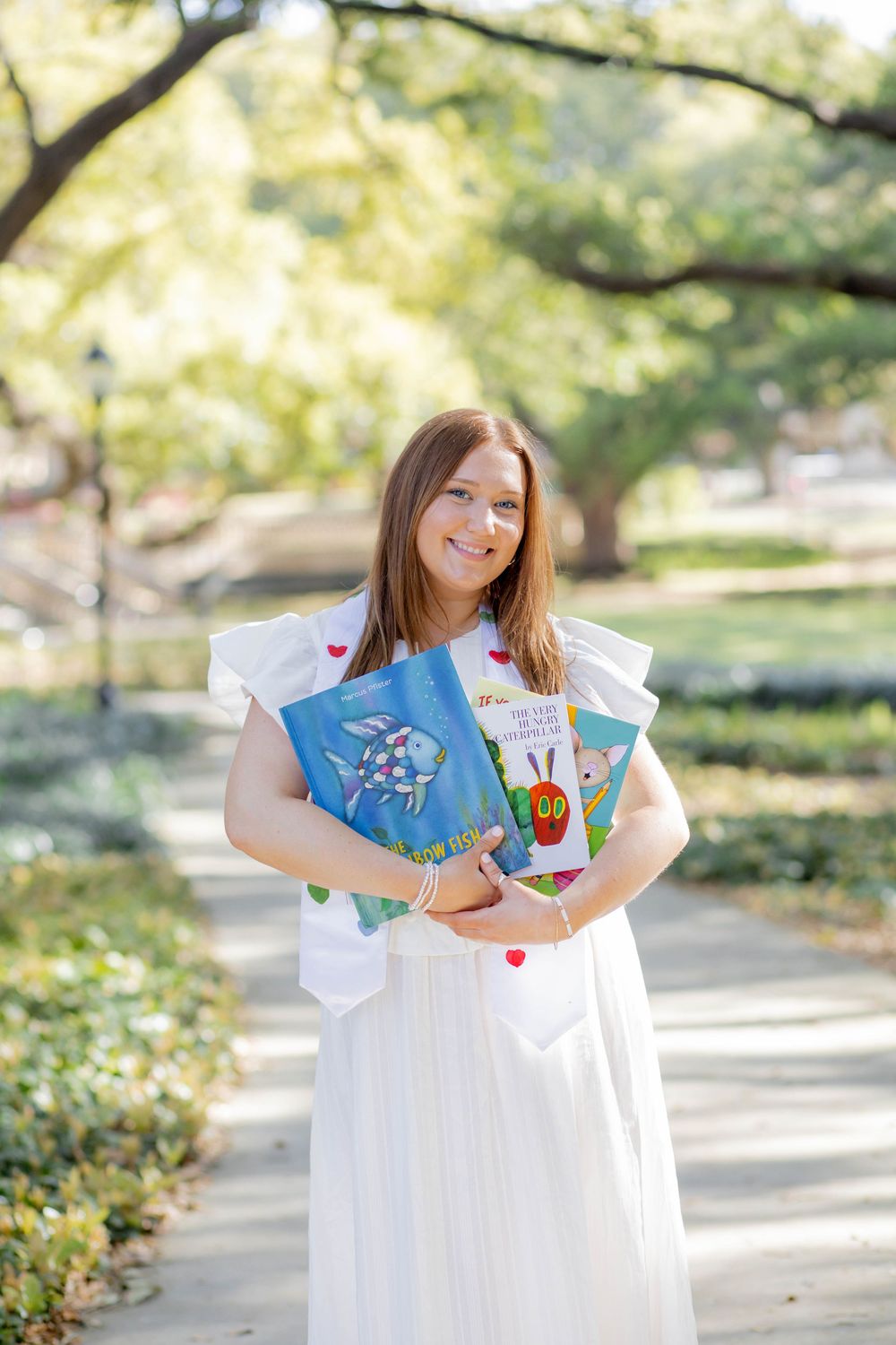 Person in a white dress holds children's picture books while standing on a sunlit garden path among green foliage.