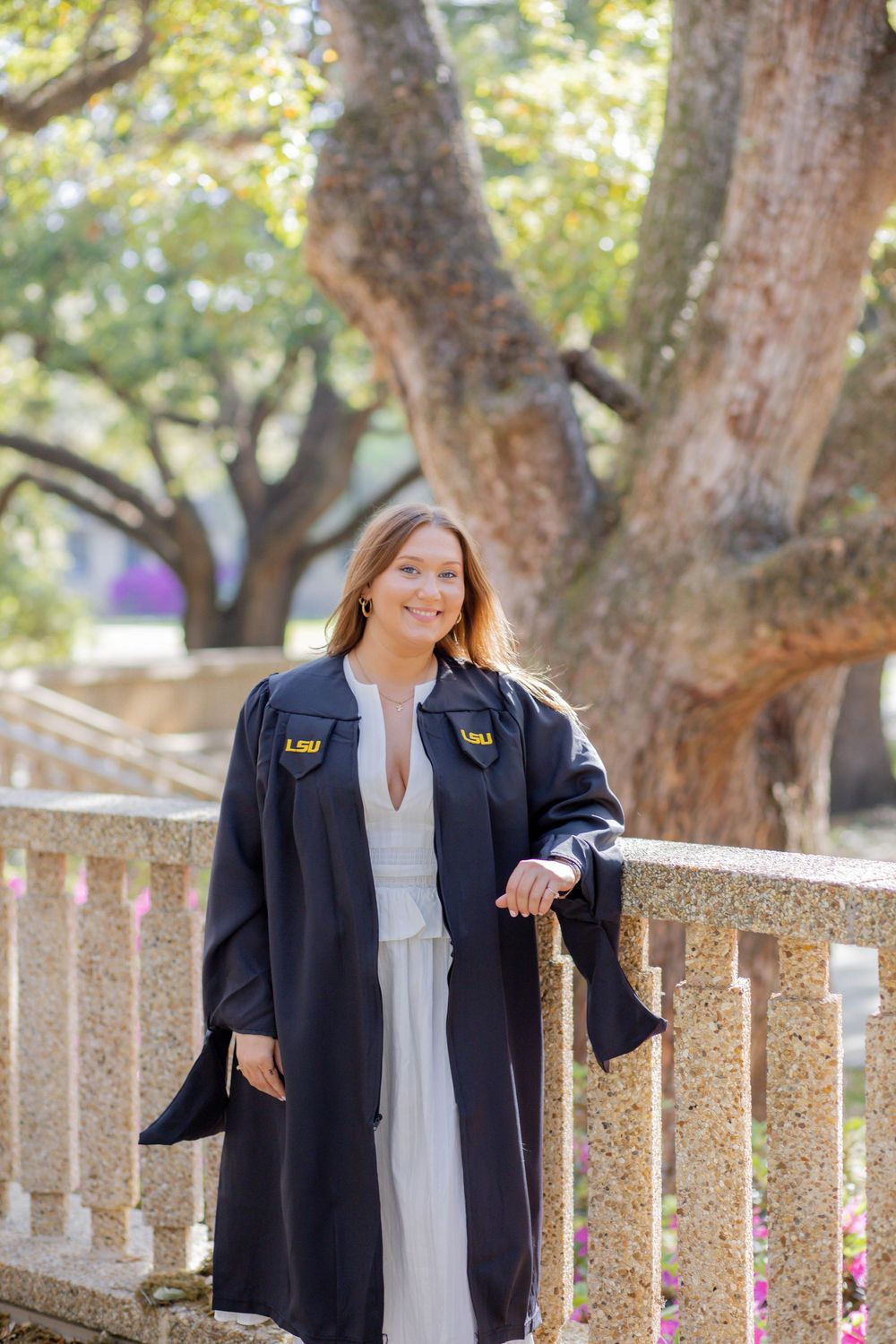 Graduate wearing a black gown and white dress poses outdoors against a blurred tree background.