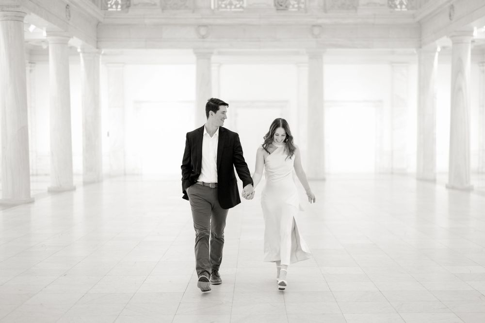 Black and white photo of a couple walking through an ornate white marble hall.