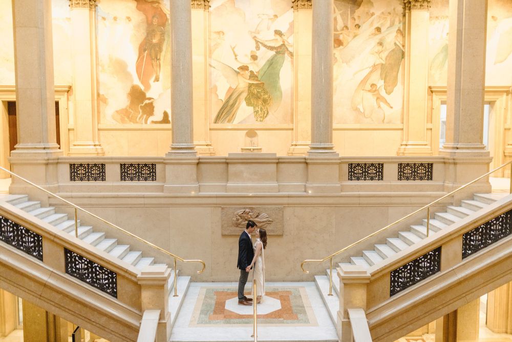 Couple embracing on grand marble staircase with classical murals in background.