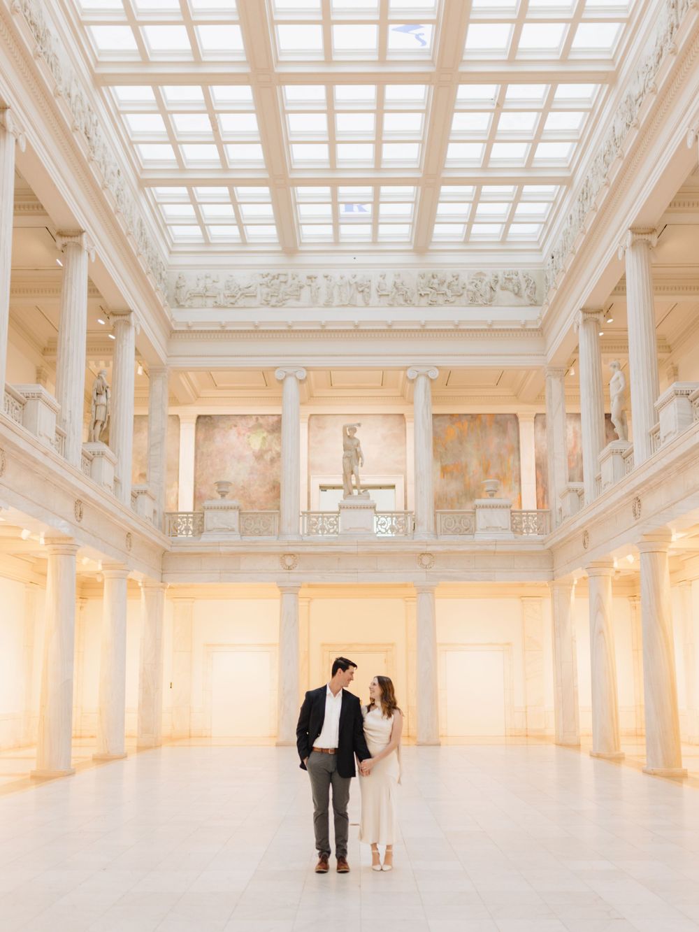 Couple photographed in an elegant white marble hall with skylight and classical details.
