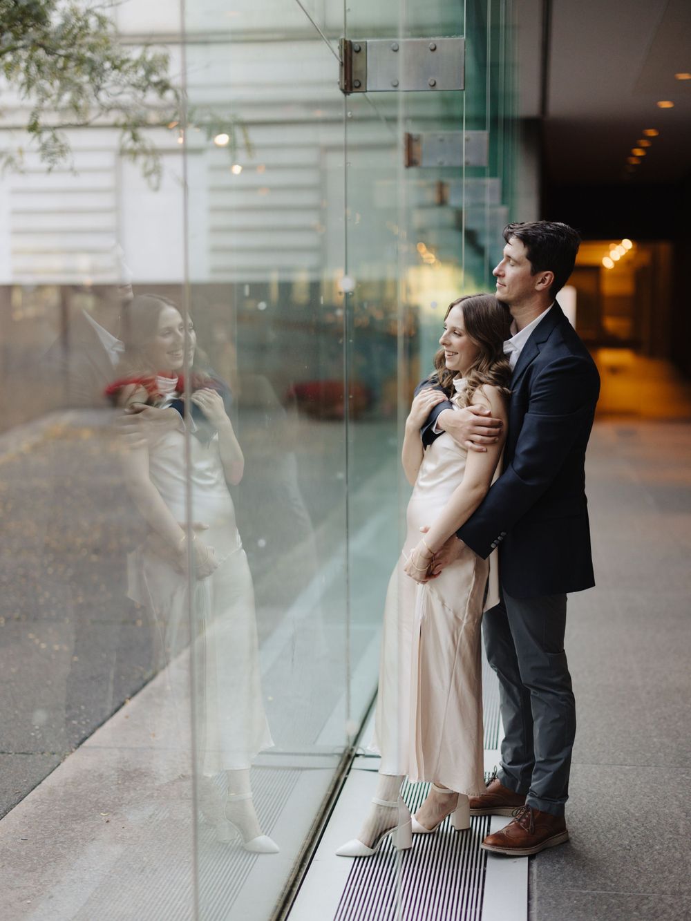 A romantic couple in formal attire embrace by a glass wall in an indoor venue at dusk.