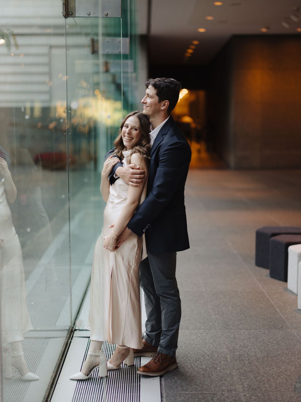 Couple shares an intimate moment in formal wear against a modern glass wall interior.