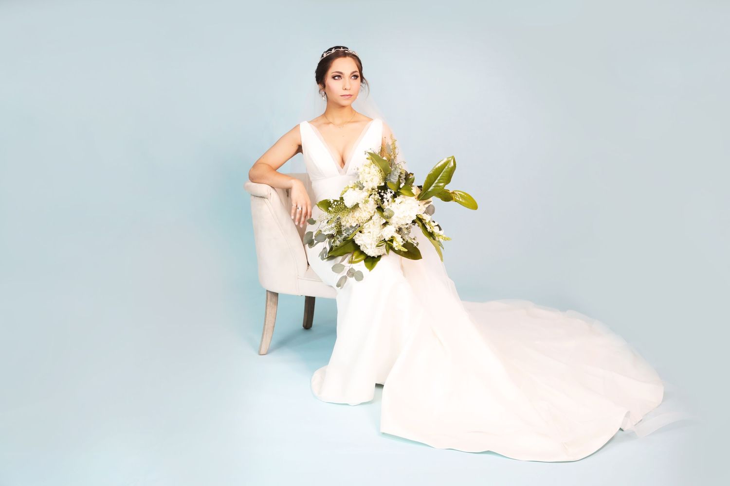 A bride in a flowing white gown poses elegantly with a bouquet of white lilies on a light blue backdrop.