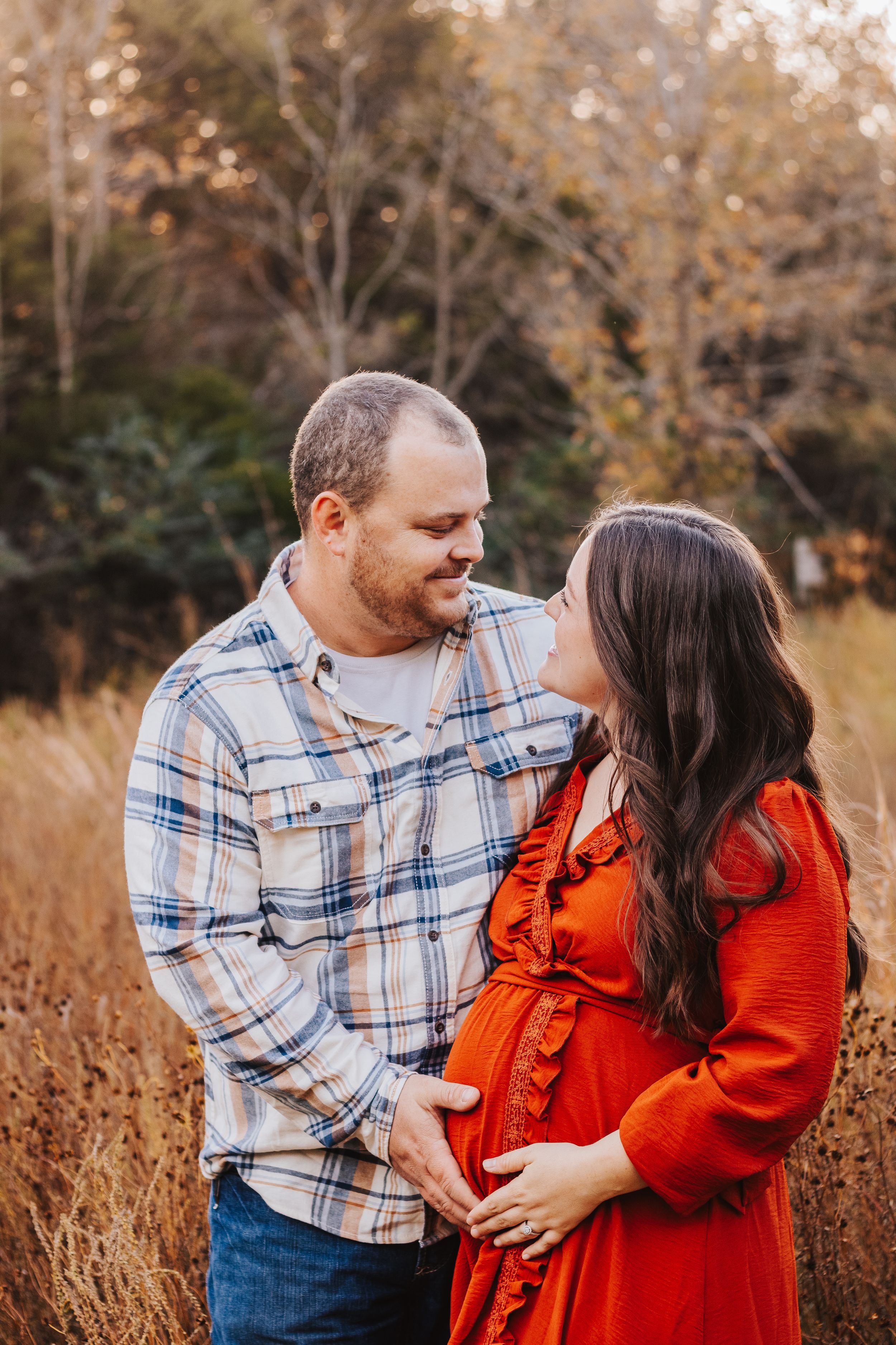Intimate maternity photoshoot with couple in plaid shirt and orange dress among fall foliage.
