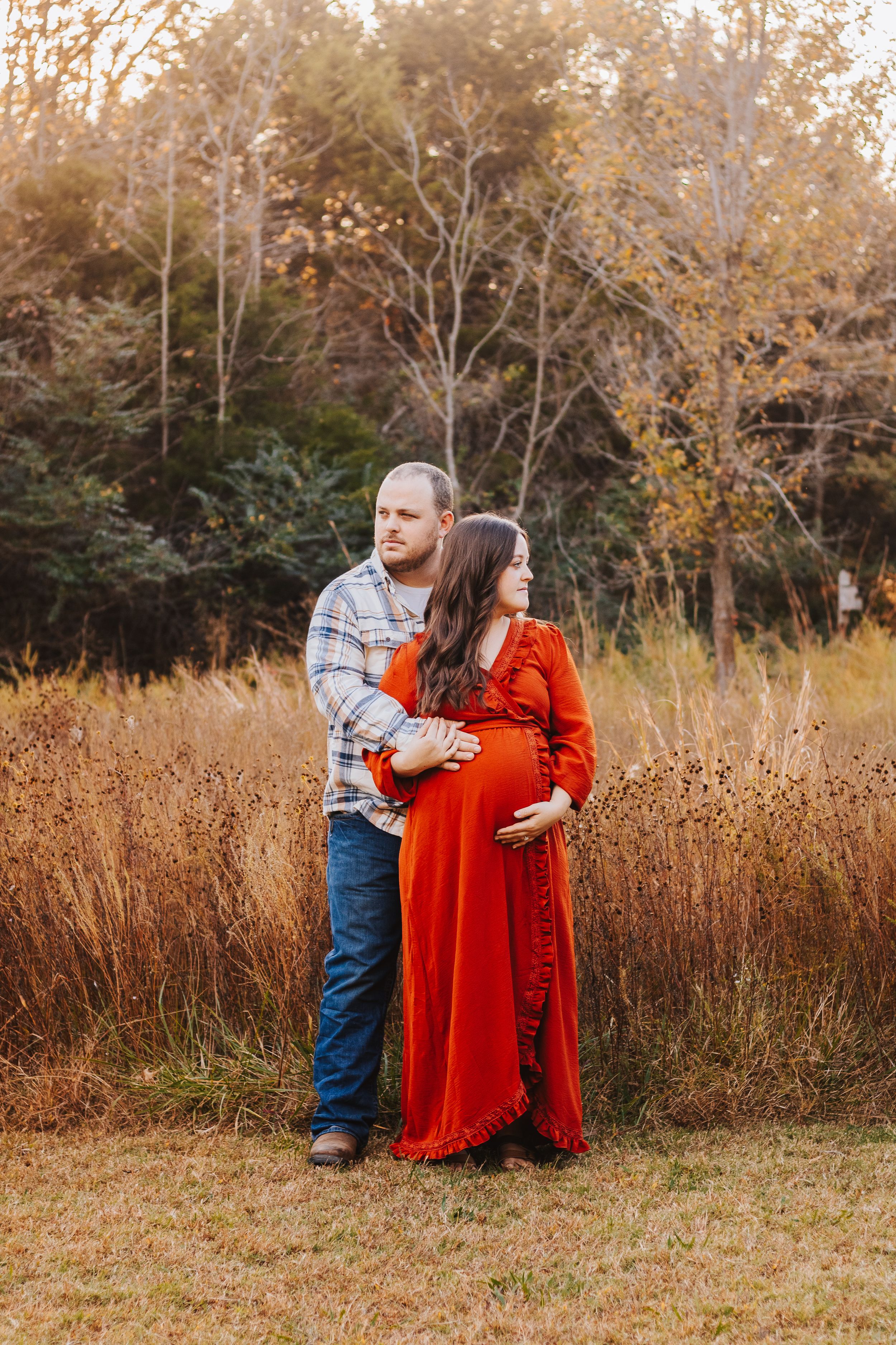 Couple poses together in field of tall brown grass during autumn sunset.