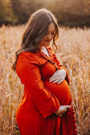 Maternity portraits in bright orange dress among autumn grasses at sunset.