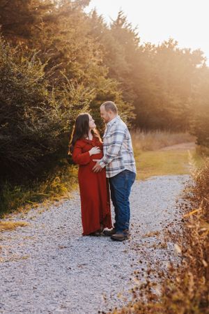 Couple stands together on gravel path during sunset maternity photo session.
