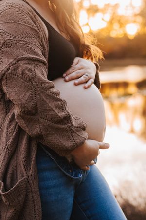 Close-up shots of pregnant belly with hands in brown sweater during sunset.
