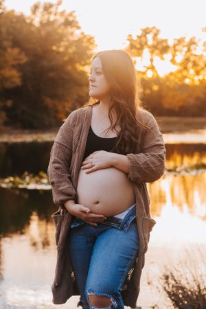 Maternity photos by lake showcase pregnant belly in brown cardigan at sunset.