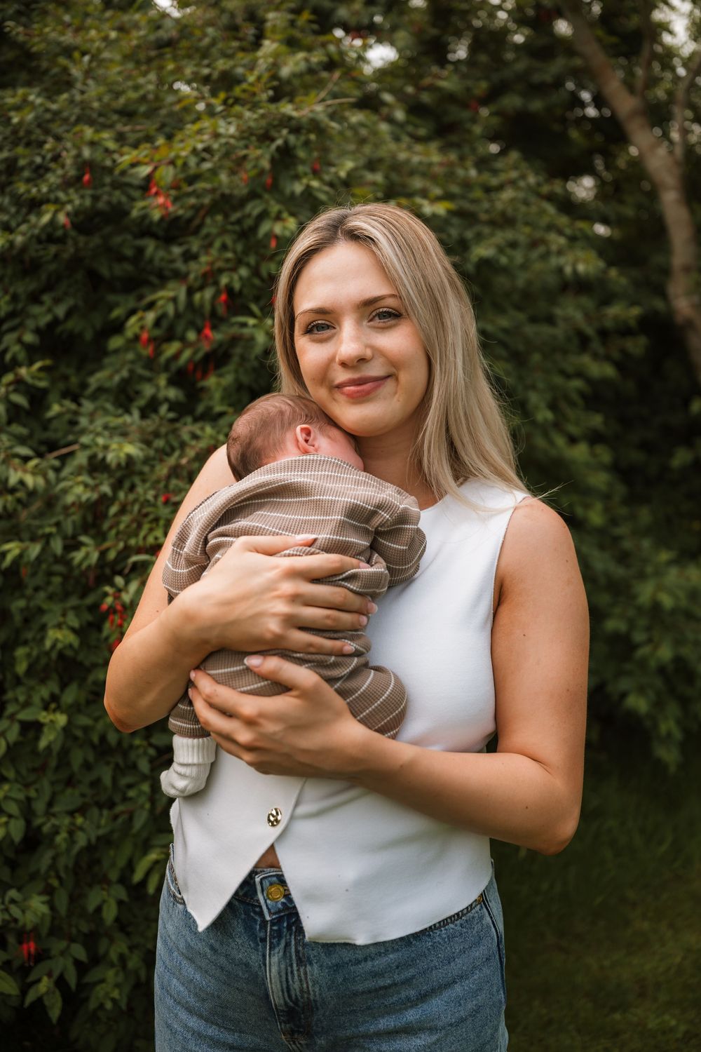 A smiling blonde woman in a white top and jeans holds a sleeping newborn baby outdoors near lush green trees.