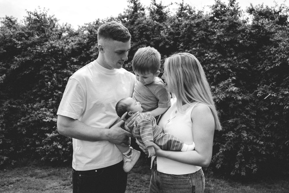 Black and white family portrait of parents holding a newborn and toddler outdoors near lush green trees.