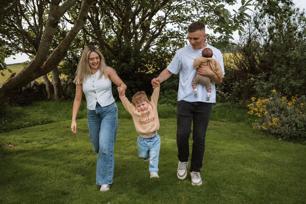 A happy family of four walks on green grass, parents swinging toddler between them while dad holds a baby.