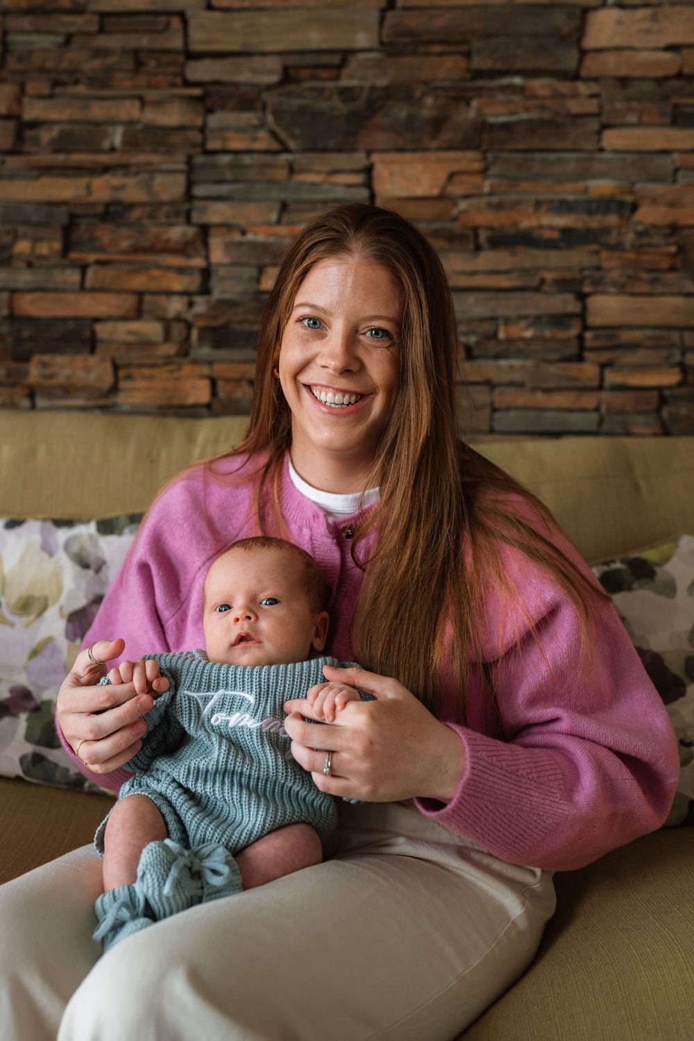 Smiling young woman with long red hair in pink sweater holding a newborn baby on a couch.