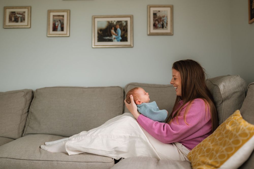 A smiling mother sits on a gray sofa cradling her newborn baby, with family photos displayed on the wall behind her.