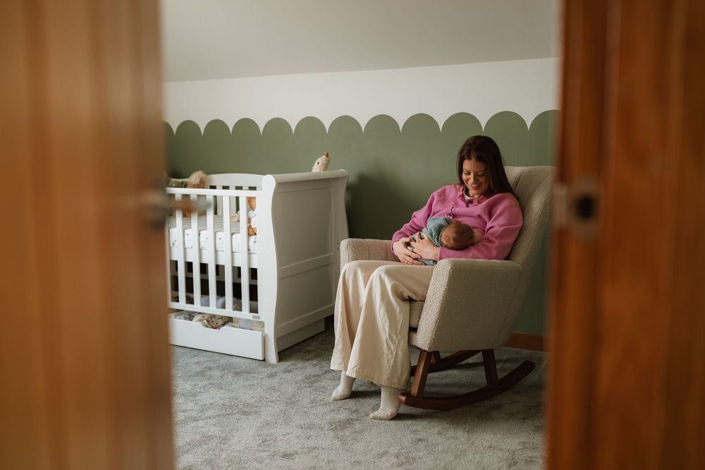 Mother sitting in a rocking chair breastfeeding her newborn baby in a green and white nursery room.