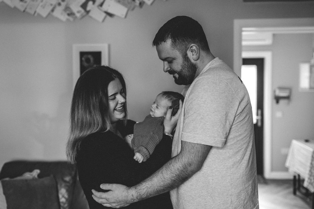 Black and white photo of a couple smiling at their newborn baby in a cozy home setting.
