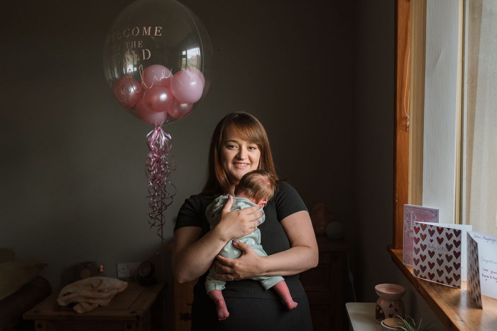 A person in a dark room holds a small baby while sitting near a window with natural light streaming in.