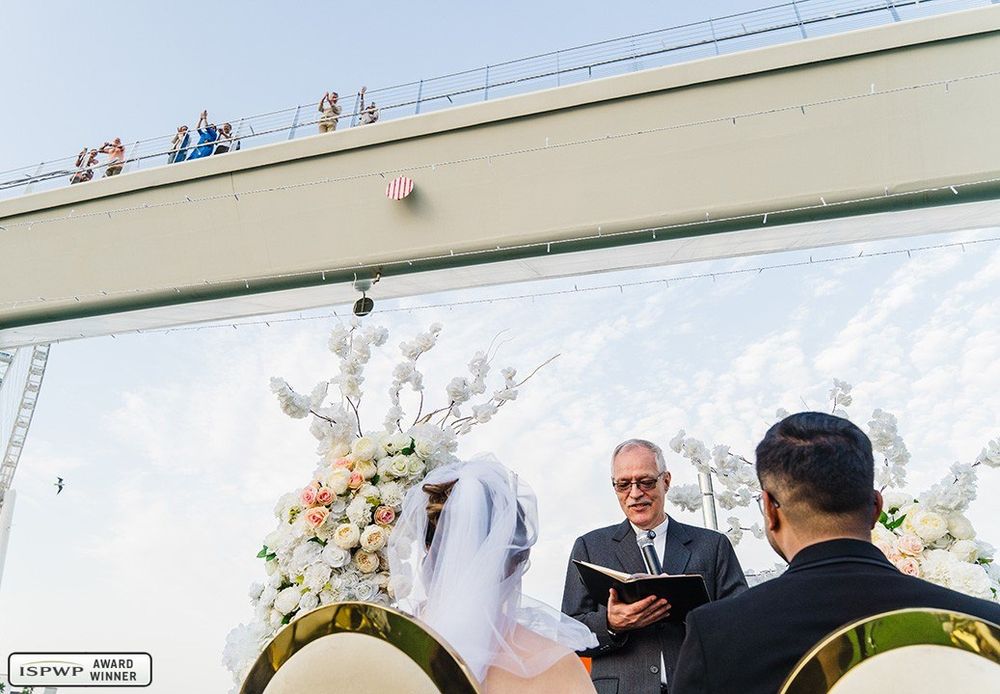 An outdoor wedding ceremony held beneath a large white-painted wall with floral decorations and officiants under a sunny sky.