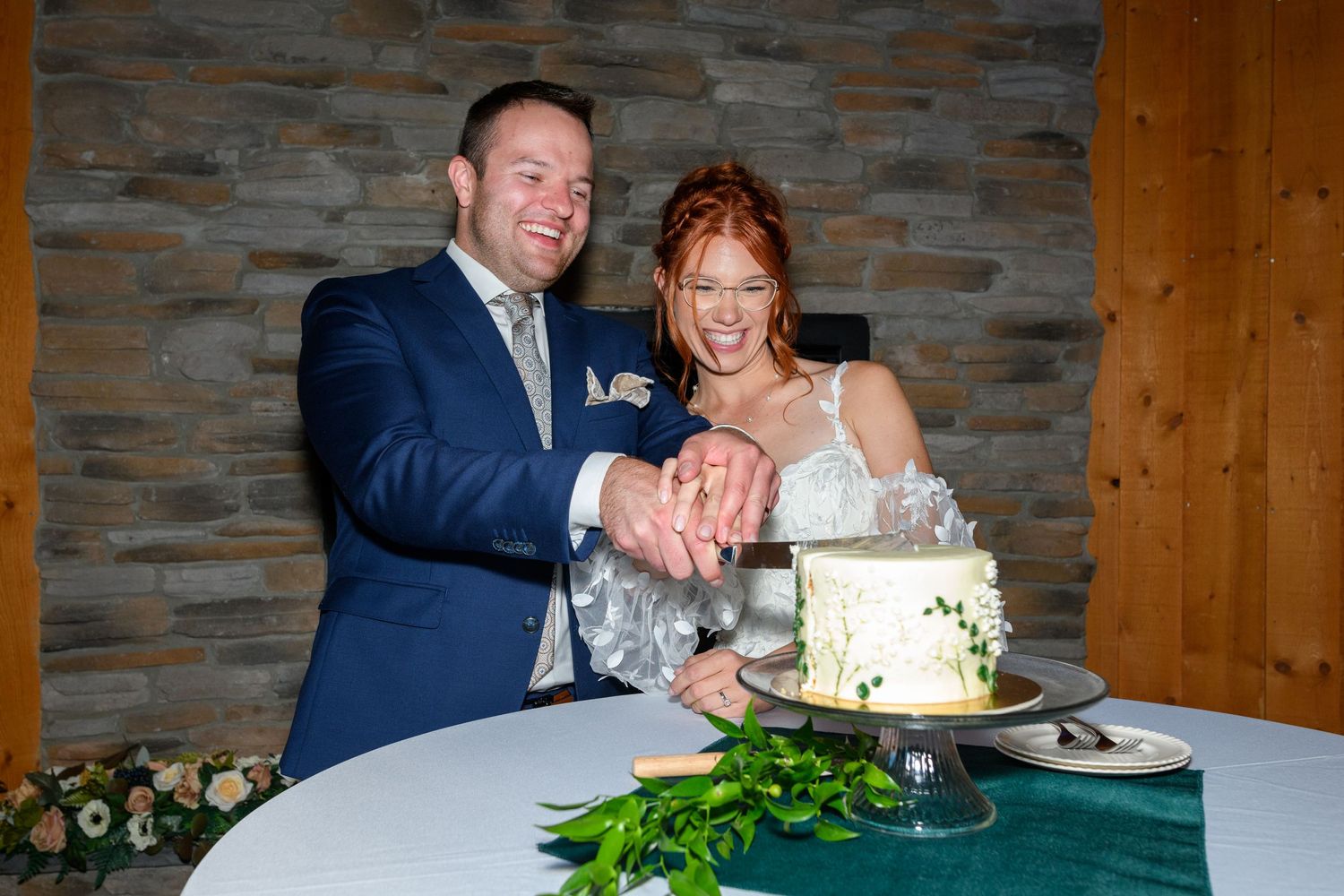 A couple cuts into their white wedding cake together at a rustic venue with stone walls and greenery decor.