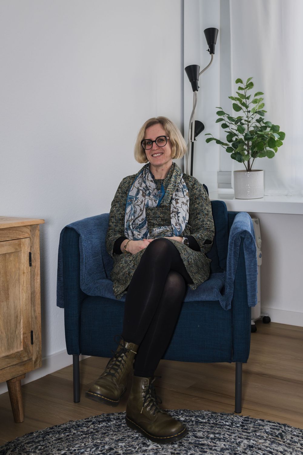 Someone relaxing in a blue armchair wearing boots and scarf, with indoor plant and lamp in background.