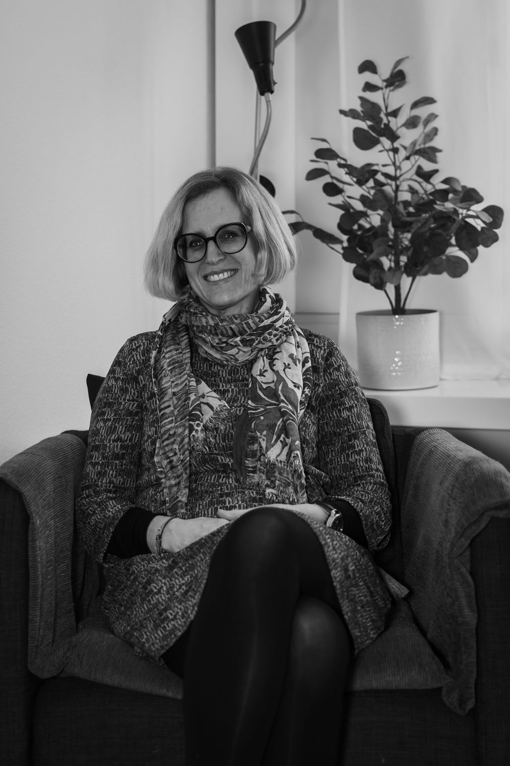 Black and white portrait of someone seated in an armchair wearing glasses, a patterned sweater and scarf near a potted plant.