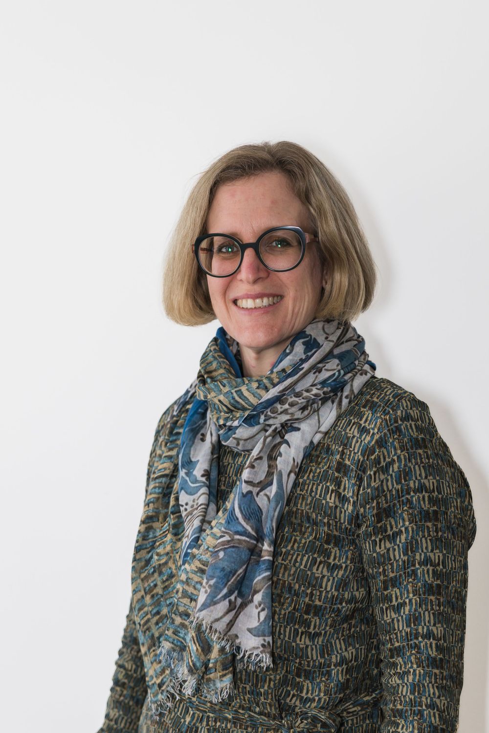 Professional portrait of someone wearing glasses, a patterned scarf and textured sweater against white background.