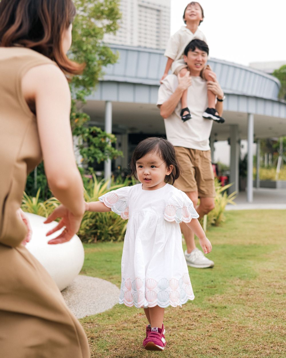 Young child in white dress running on grass with family in background.