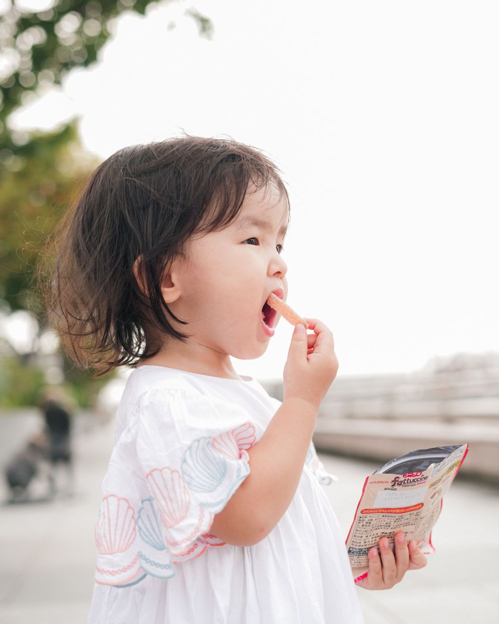Small child in white dress eating snack while holding package outdoors.