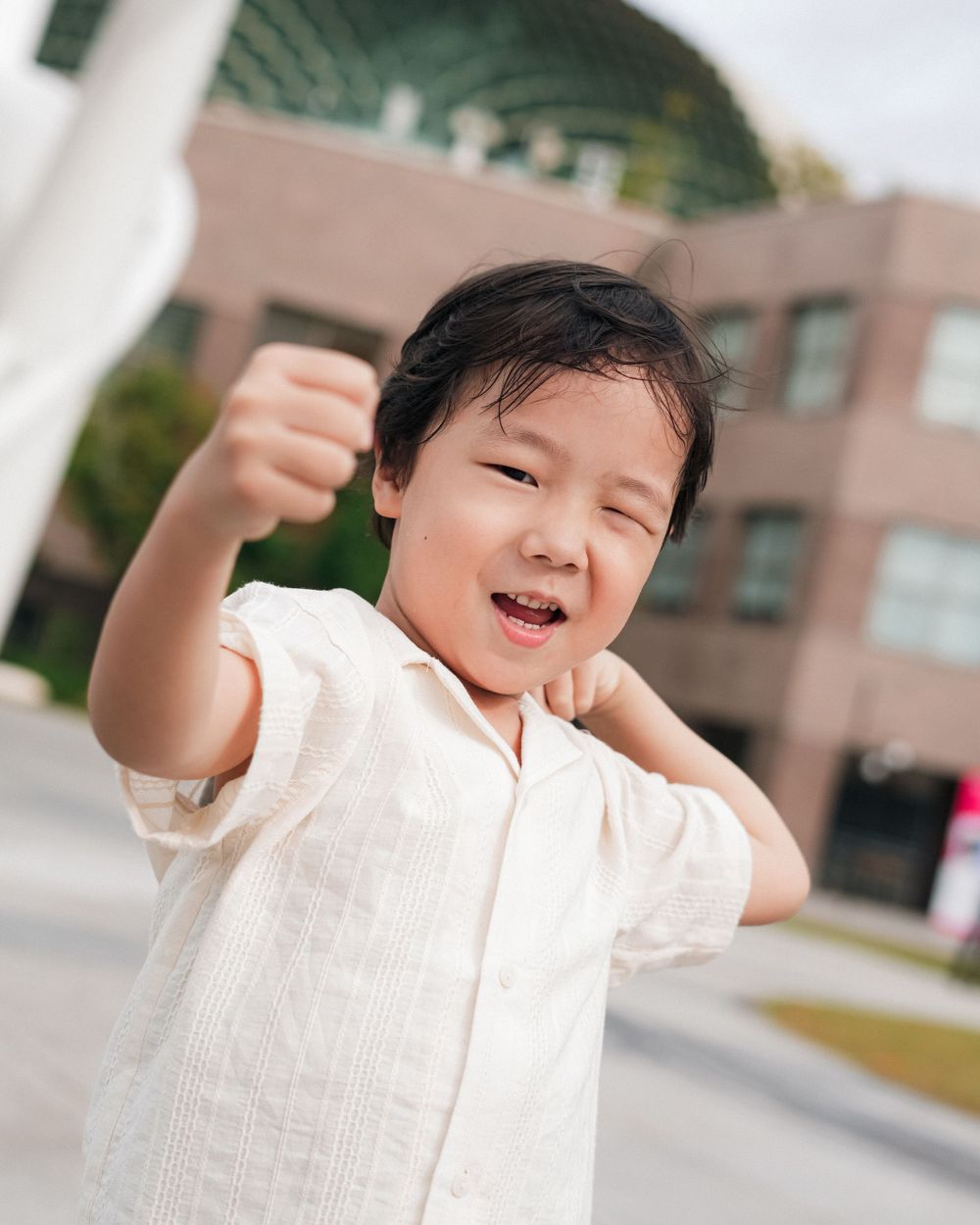 A child in a white shirt smiles and raises their fist joyfully outdoors.