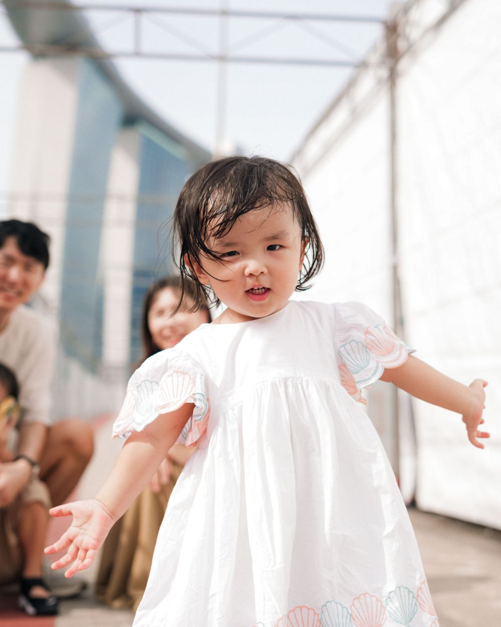 A small child in a white dress gestures expressively against a bright background.