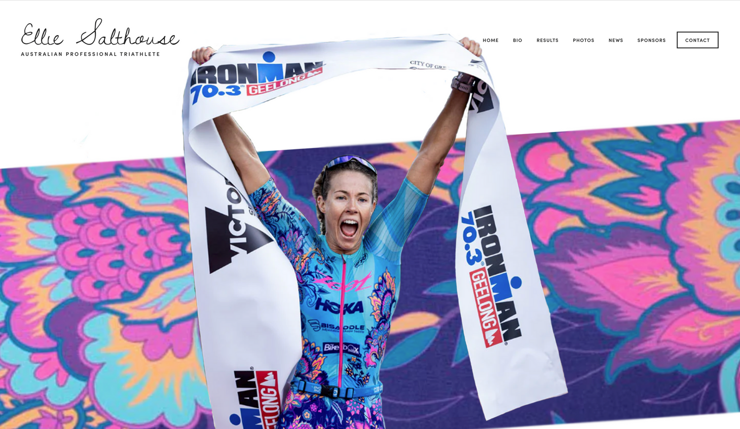 An athlete celebrates victoriously with raised arms while wearing a blue and black triathlon outfit against a vibrant pink backdrop.