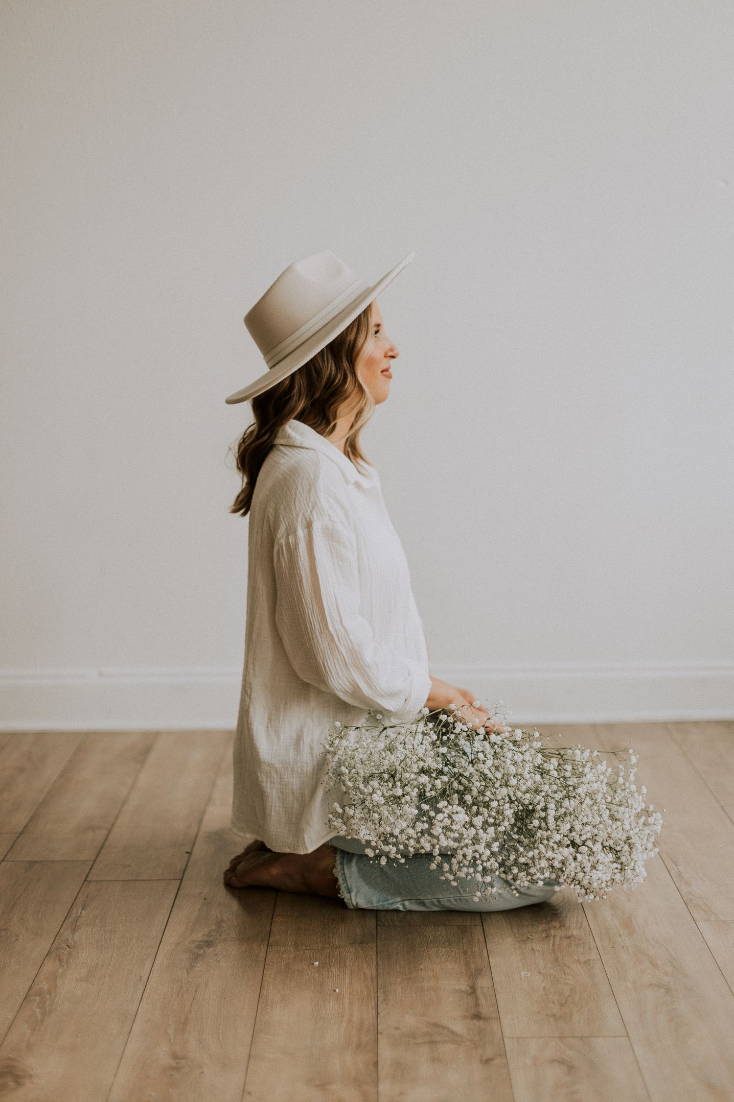 Side profile of person in white shirt and hat sitting with dried flowers on wooden floor.