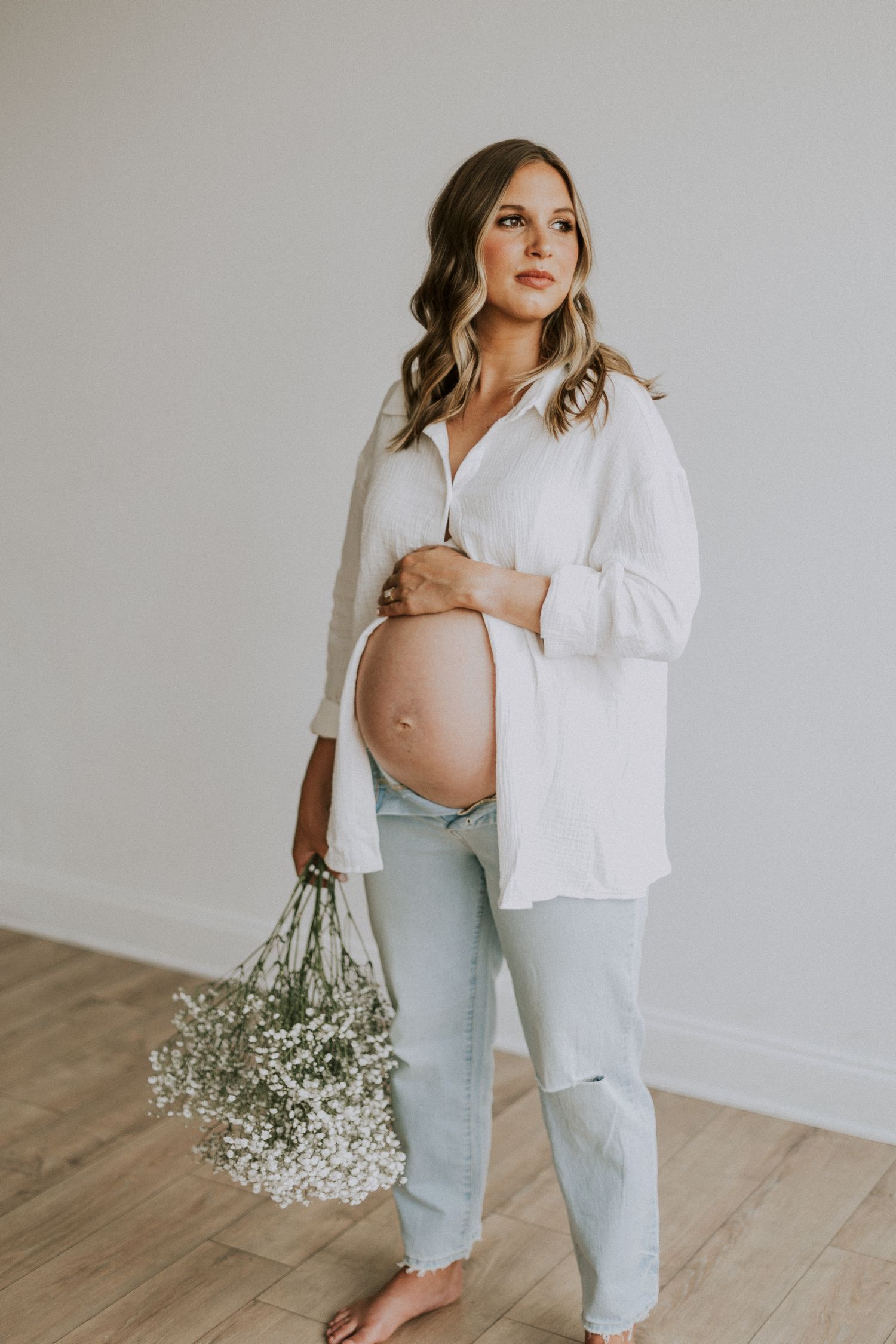 Expectant parent in white shirt holding belly while posing with baby's breath flowers.