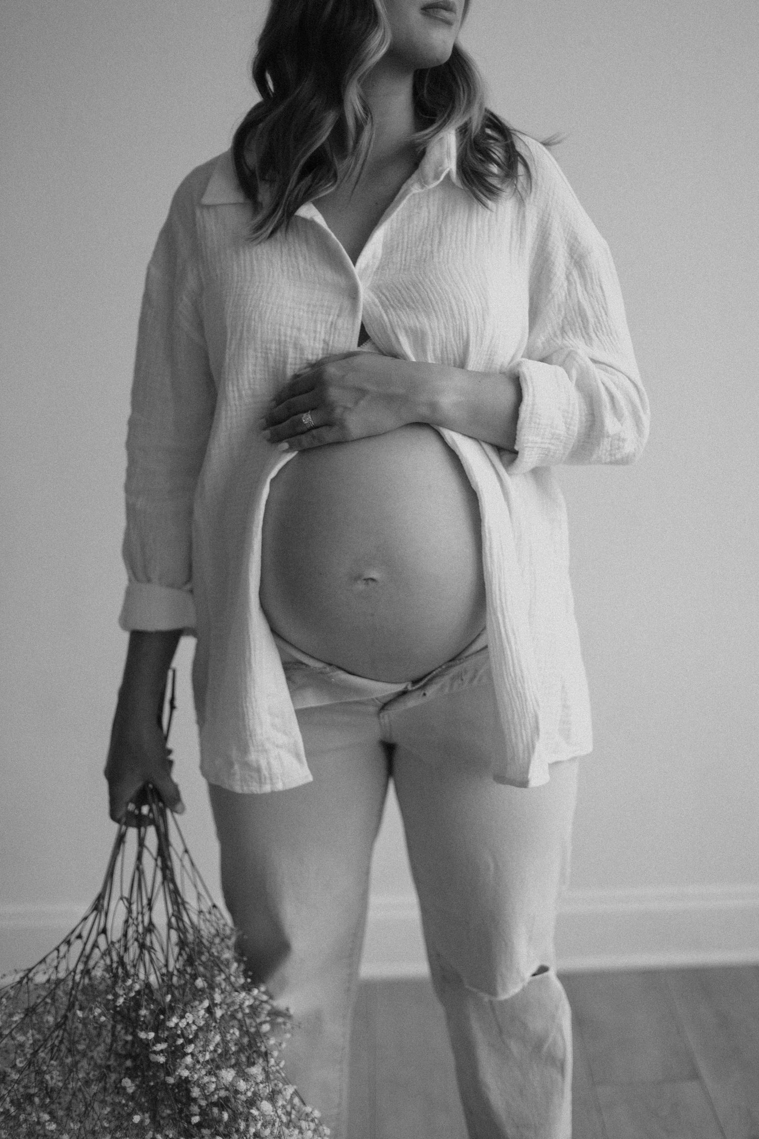 Maternity photo showing pregnant belly in white button-down shirt and casual wear holding dried flowers.