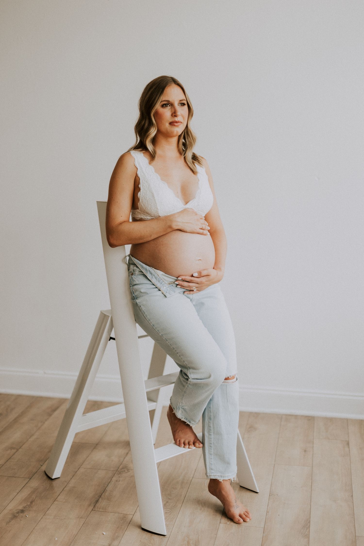 Maternity portrait with person sitting on white stool wearing ripped jeans and white top.