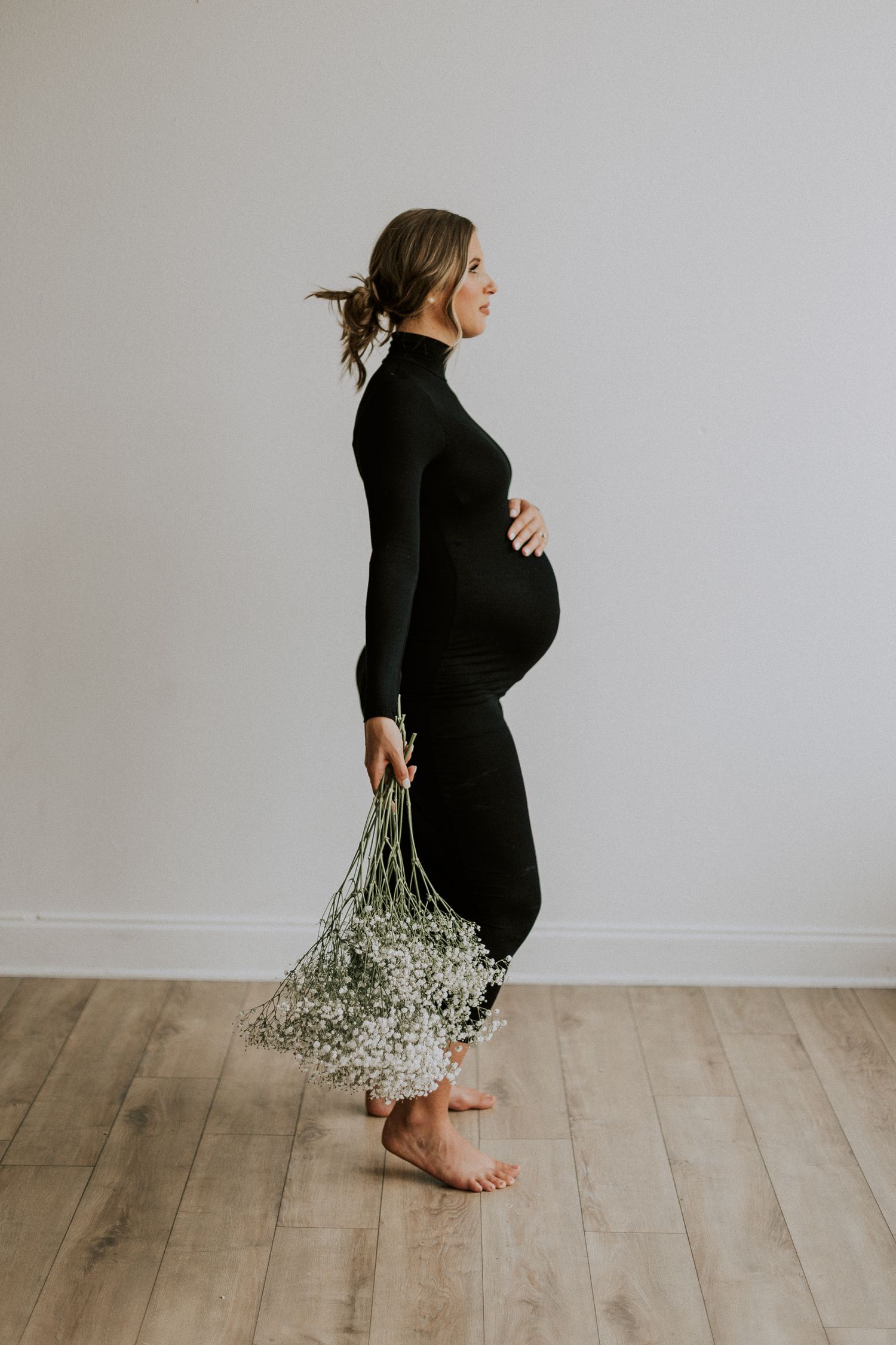 Side view of an expectant mother in a black dress holding dried flowers against a minimalist backdrop.