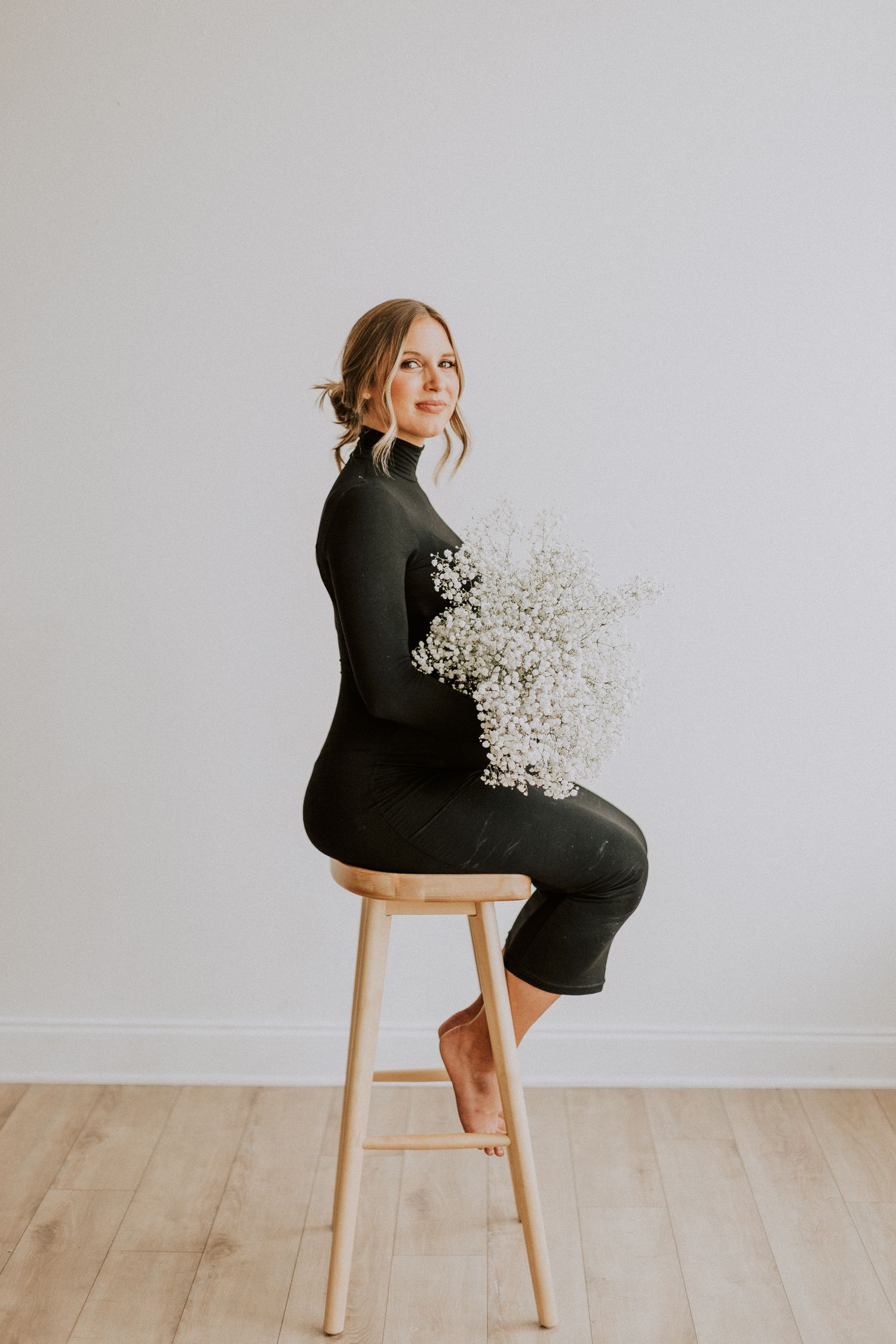 Maternity portrait featuring a woman in a black dress holding baby's breath flowers while seated on a wooden stool.