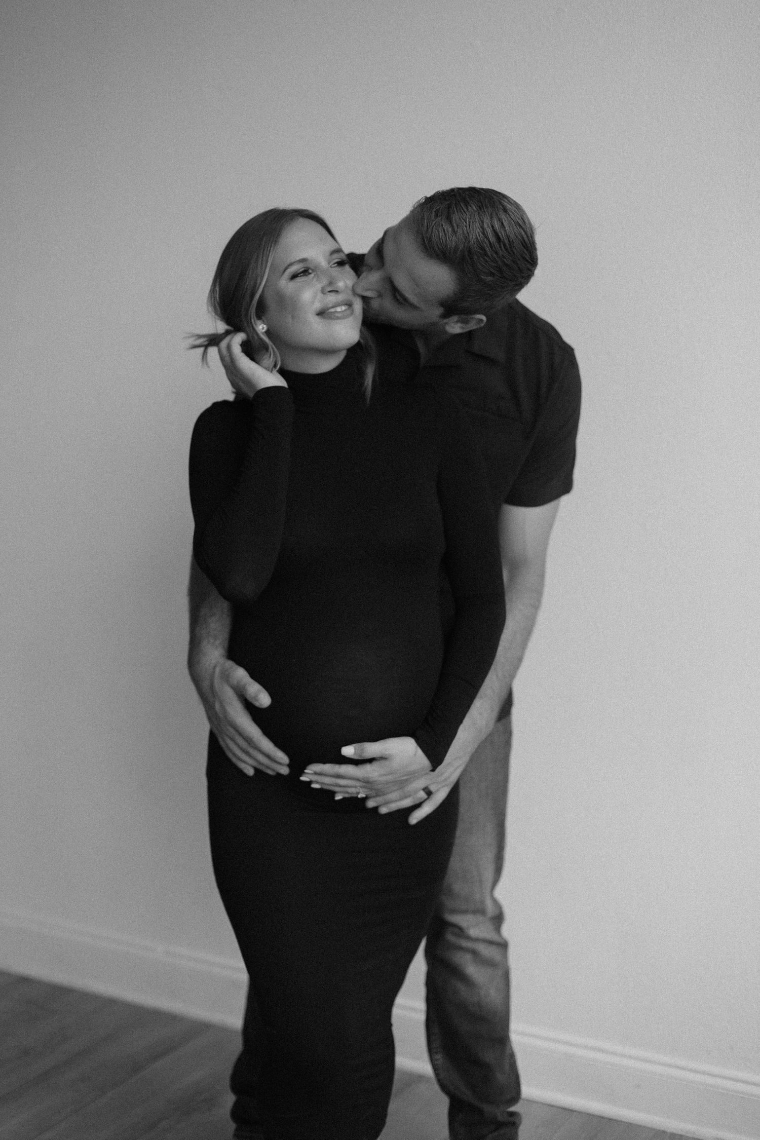 A tender maternity photo shows an intimate moment between a couple in black clothing against a light background.