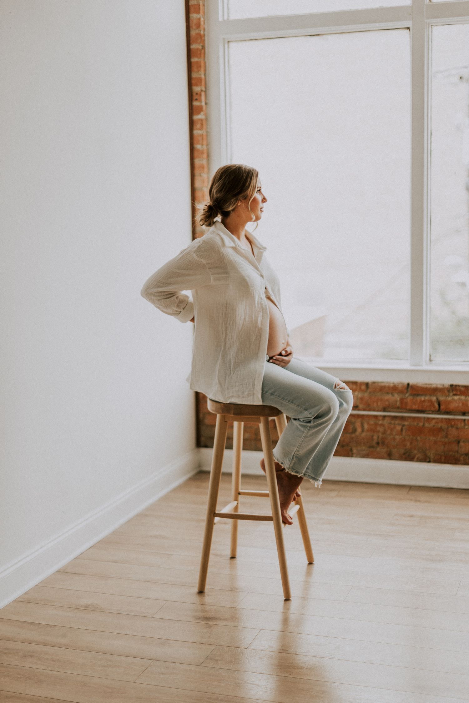 Person in white button-down shirt sitting on wooden stool by window during maternity photoshoot.