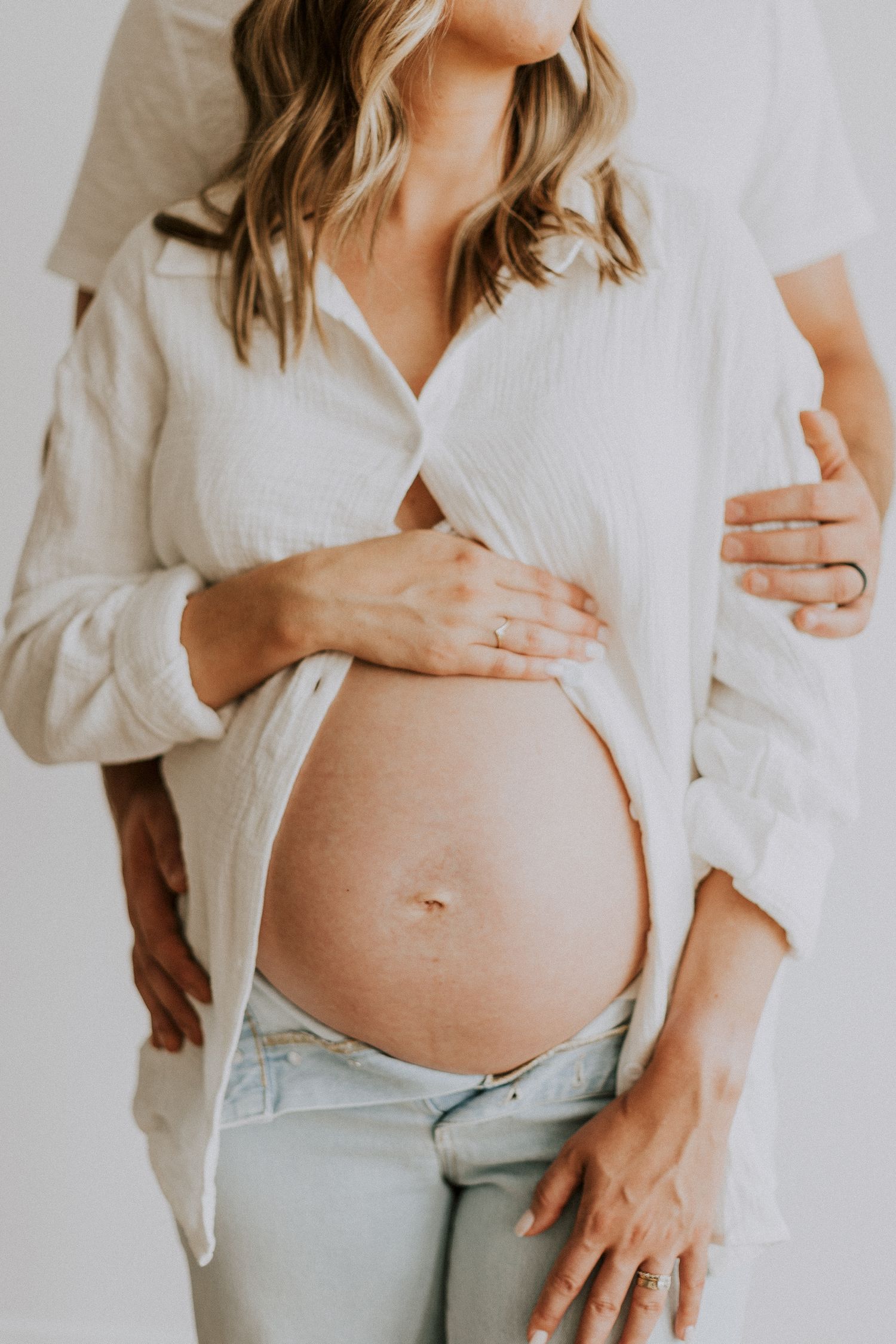 Close-up maternity photo showing pregnant belly with white button-down shirt and light jeans.