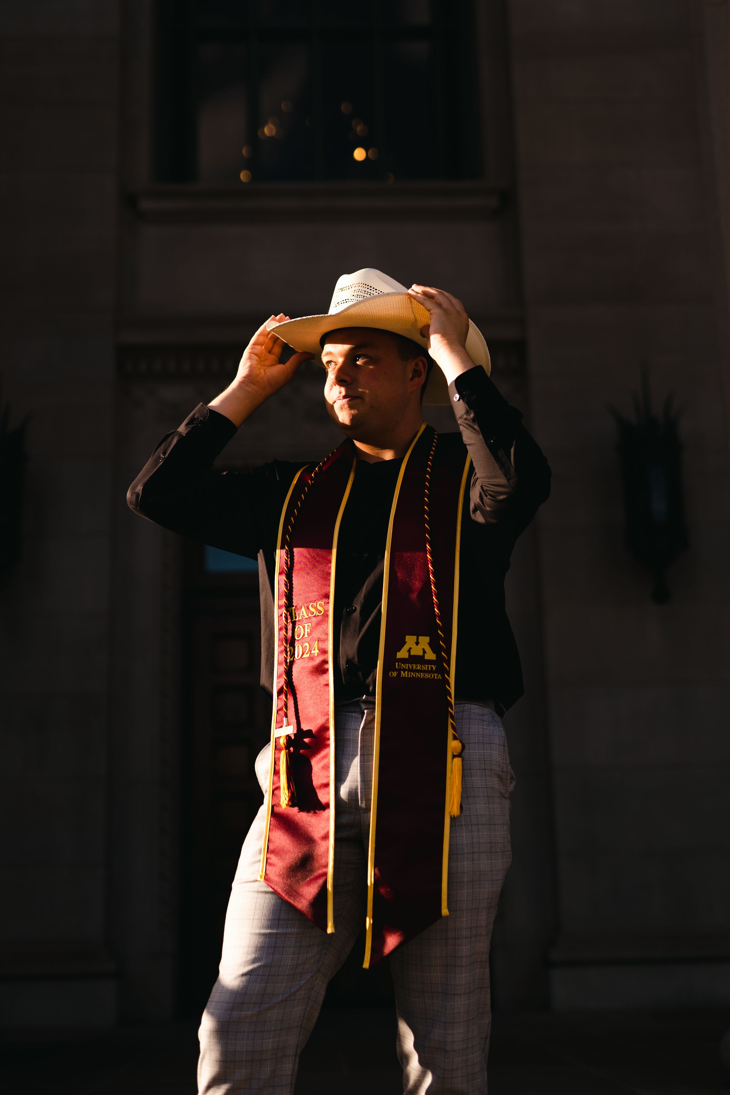 Graduate adjusts graduation cap while wearing ceremonial stole in dramatic lighting.