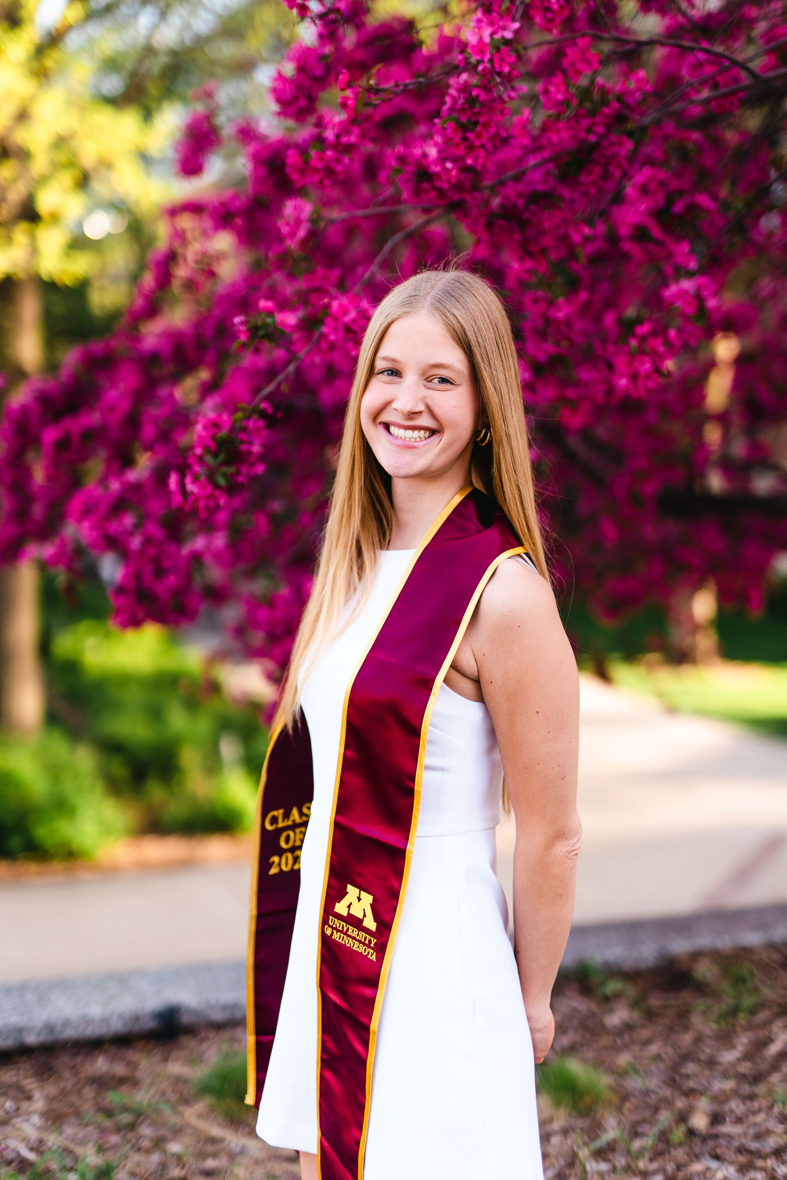 Graduate wearing white dress and maroon sash poses in front of blooming magenta flowers.
