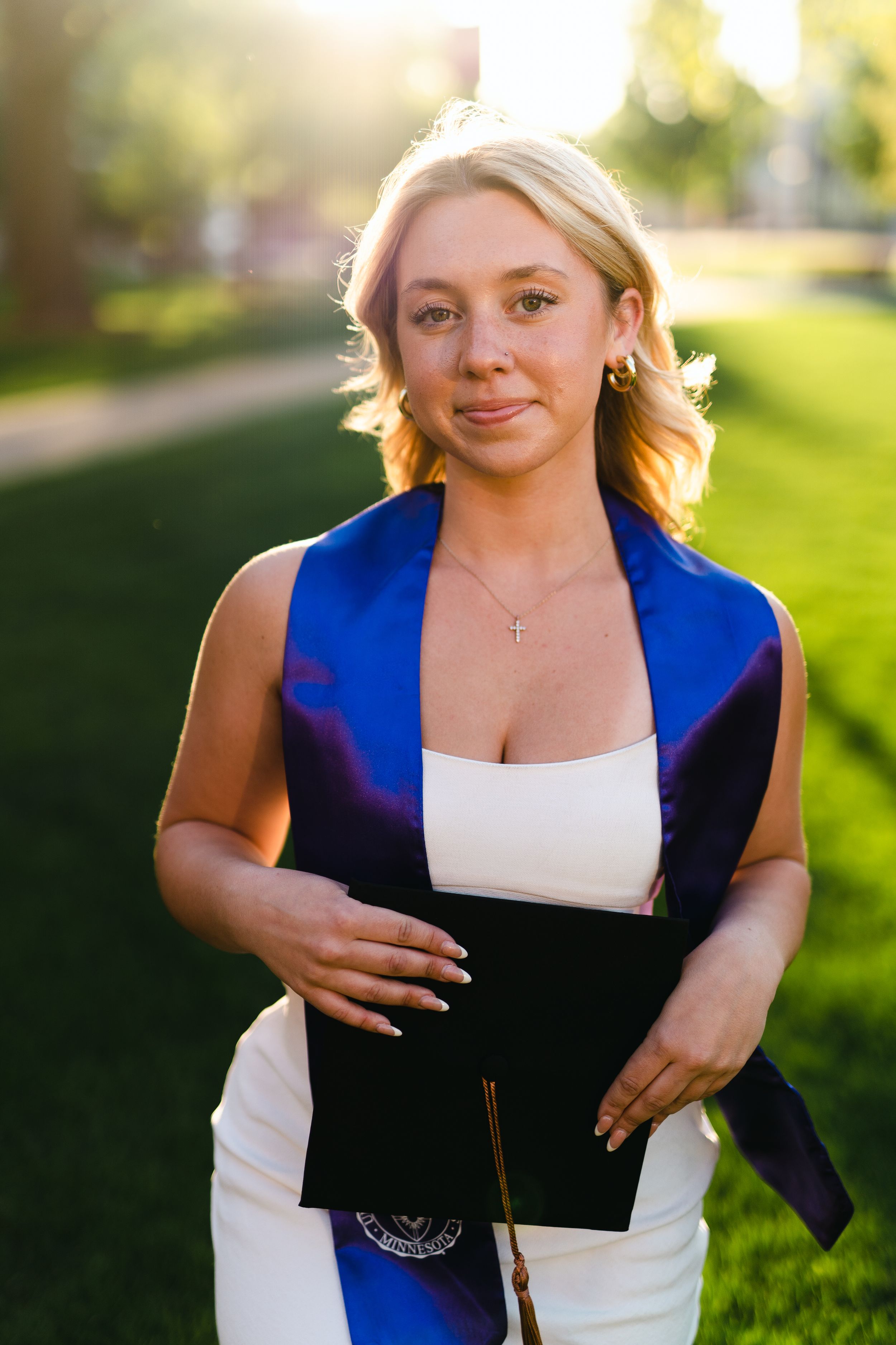 Graduate in white dress and blue stole poses outdoors during golden hour.