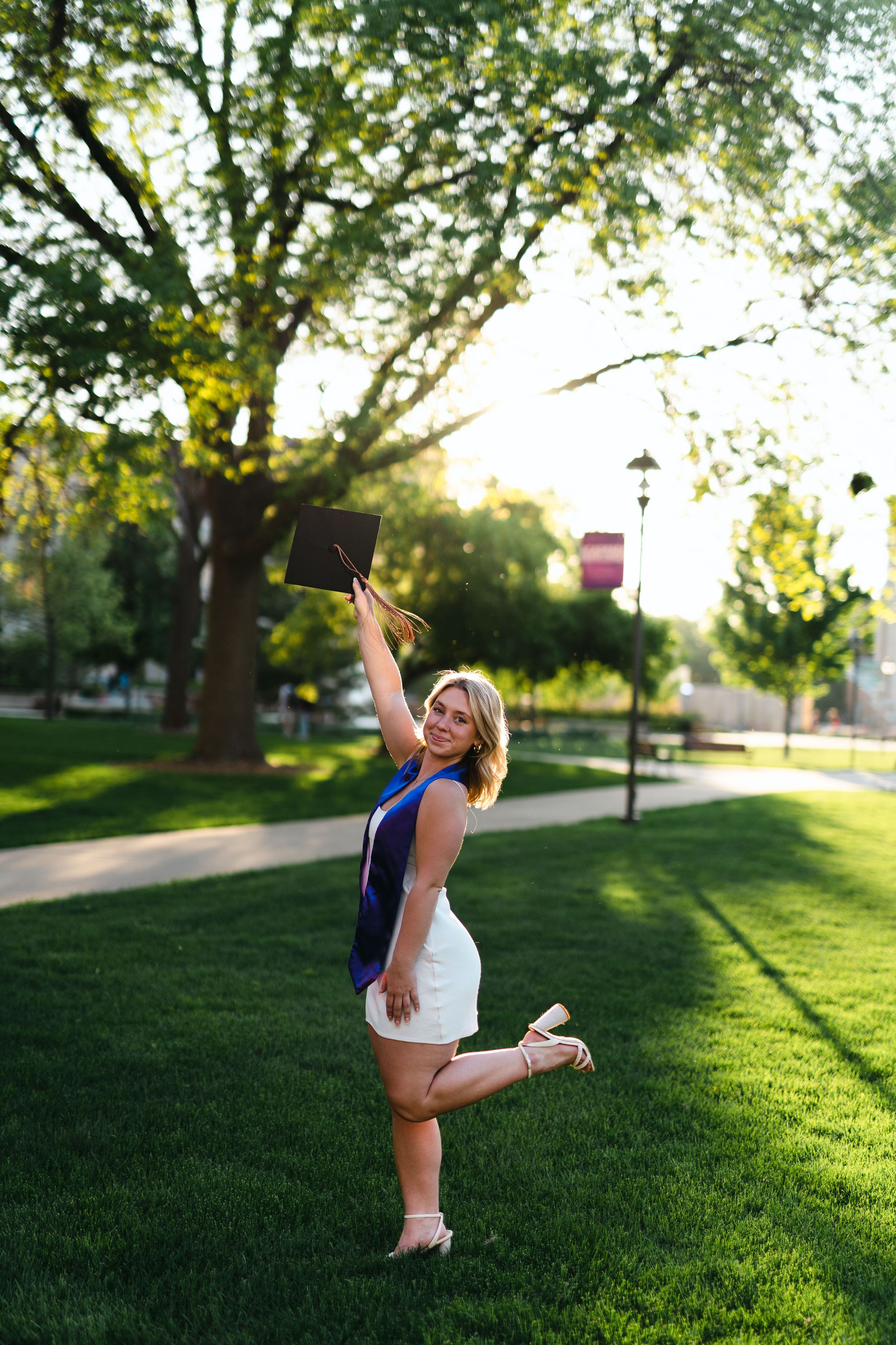 Graduate in white dress celebrates outdoors by raising diploma.