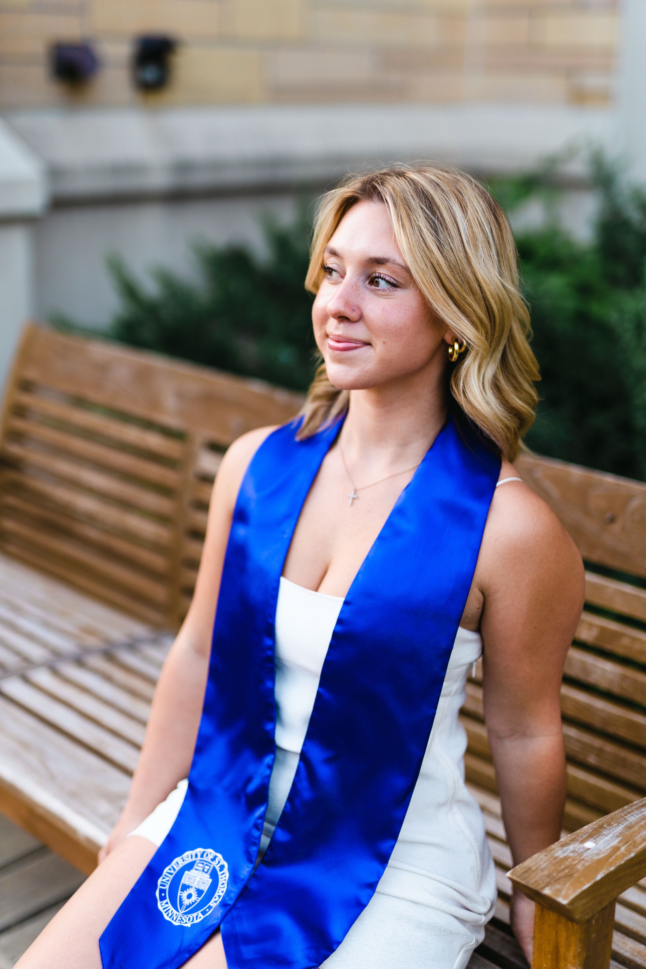 Graduate in blue stole sits on wooden bench outdoors.