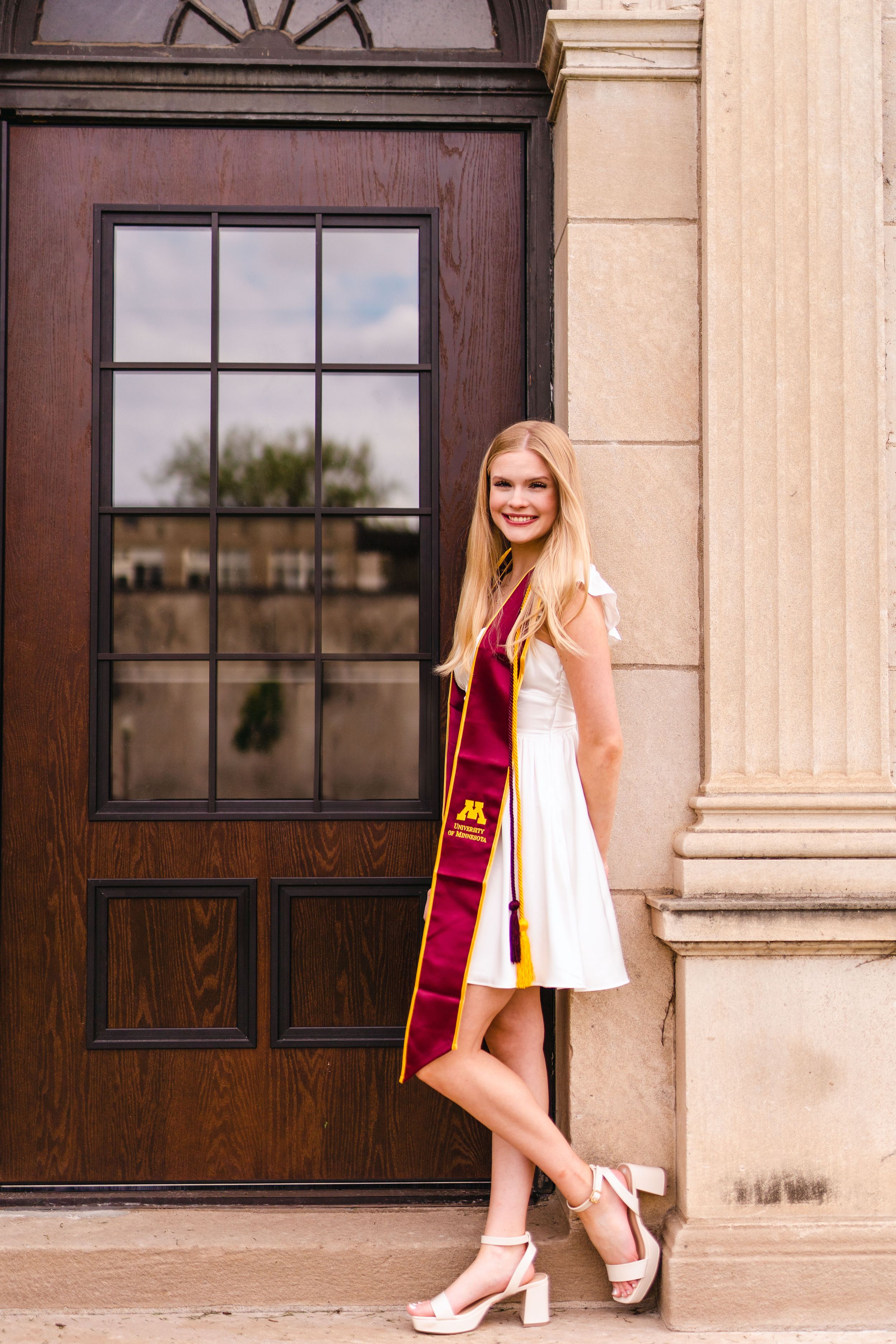 Graduate in white dress poses against classic architectural doorway.
